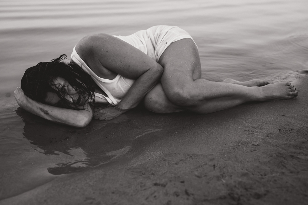 Frau liegt zusammengerollt im seichten Wasser am Sandstrand, Schwarz-Weiß – kraftvolle Seelenfotografie von MALEA Fotografie